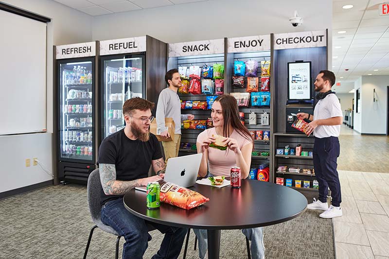 Two people sit eating snacks and working at a table in front of a micro market where two other people converse over snack items.