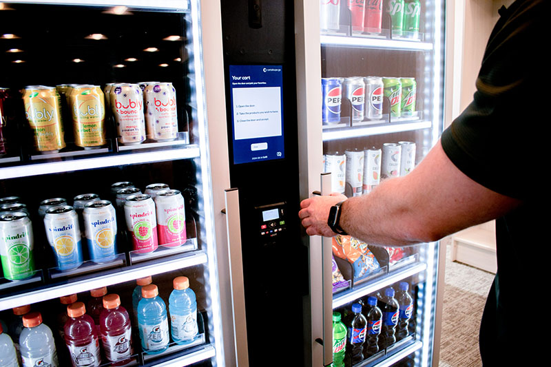 Customer reaches for door handle of Smart Store cooler packed with drinks and snacks.